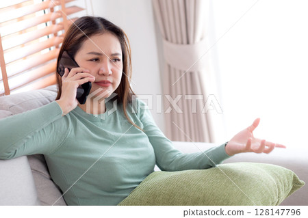 Asian young woman stress and worried sitting on sofa while speaking on smart phone in living room. 128147796