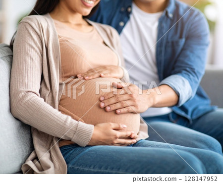 一對新婚夫婦中的一位年輕亞洲男子輕輕撫摸著坐在沙發上的孕婦的肚子 128147952