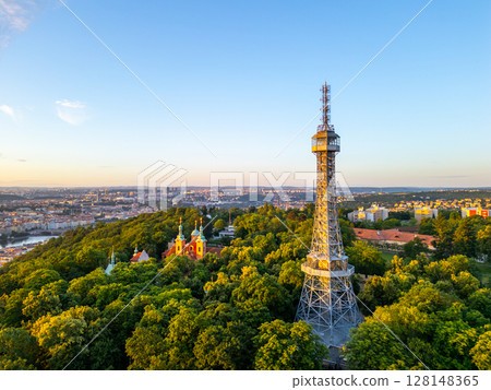 The Petrin Lookout Tower stands tall against the early morning light, surrounded by lush greenery. A picturesque view of Prague unfolds as the city awakens at dawn. 128148365