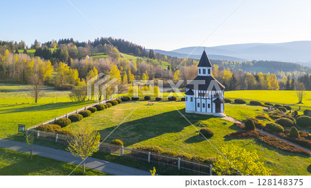 The white octagonal Tesarov Chapel stands gracefully in Korenov, surrounded by lush greenery and gentle hills in the Jizera Mountains during a sunny day. 128148375