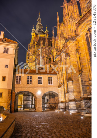 The illuminated Covered Bridge links architectural marvels at Prague Castle, highlighting the intricate details of Saint Vitus Cathedral under the night sky and street lamps. 128148376