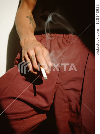 Tattooed hand holding a lit cigarette with curling smoke Tattooed hand holding a lit cigarette with curling smoke 128148938