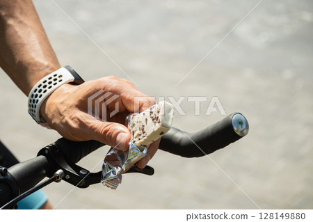 Energy or protein bar in cyclist hand. Man snacks and riding bike. Healthy protein snack or energy bar for sporty lifestyle. Close up Energy or protein bar in cyclist hand. Man snacks and riding bike. Healthy protein snack or energy bar for sporty lifestyle. Close up 128149880