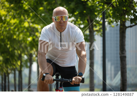 Caucasian cyclist in motion in white t-shirt rides bicycle through urban public park. Sporty man wears reflective sunglasses. Outdoor activity. 128149887