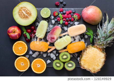 In the center on ice cubes a variety of fruit sorbet cones around ingredients fruits and berries on a black background. Variety of fruit and berries sorbet cones. Horizontal. Top view. 128149929