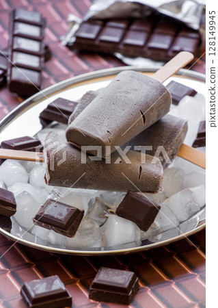 Chocolate ice cream on a stick on a silver platter with ice cubes and pieces of chocolate near pieces of chocolate on a chocolate background. Chocolate ice cream popsicles. Vertical. Close. 128149945