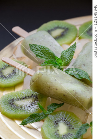 Kiwi ice cream popsicles are beautifully presented atop sliced kiwi fruits. Fresh mint leaves add a vibrant touch, enhancing the desserts visual appeal and flavor. 128149946