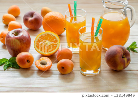 Fruit juice in glasses with ice near the jar of juice and scattered apricots, peaches, apple, orange on a light wooden background. Apricot peach apple orange juice with ice. Horizontal. 128149949