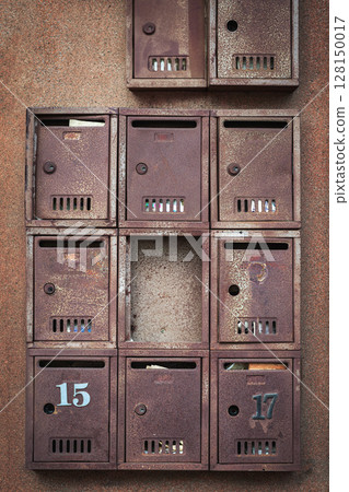 Old rusted metal mailboxes with peeling numbers and missing compartments attached to textured urban wall. Concept of urban nostalgia and vintage 128150017
