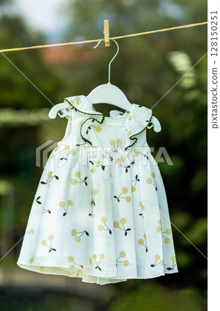 Baby clothes hanging on clothes hanger with clothespin, natural materials. Bright, cute dress adorned with floral patterns hangs from clothesline in lush garden. Spring summer style, child dress 128150251