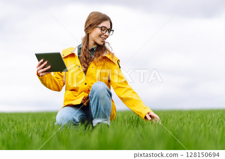 Young farmer woman in a green field, intently using a tablet. She wears a yellow jacket and glasses 128150694