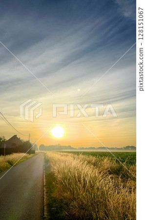 Experience the serene beauty of a vivid sunrise over a peaceful country road and fields 128151067