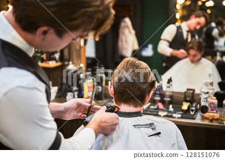 Back view of professional hairdresser making stylish haircut. Barber in fancy outfit using a comb and electric shaver to cut extra hair on nape. 128151076