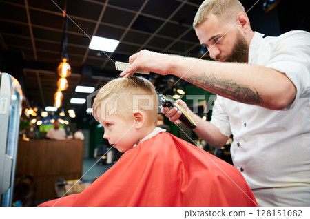 Barber using comb and shaver to cut hair. Professional hairdresser shaving little kid's nape. 128151082