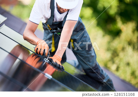 Worker building photovoltaic solar panel system on rooftop of house. Close up of man engineer in gloves installing solar module with help of hex key outdoors. Renewable energy generation concept. 128151108