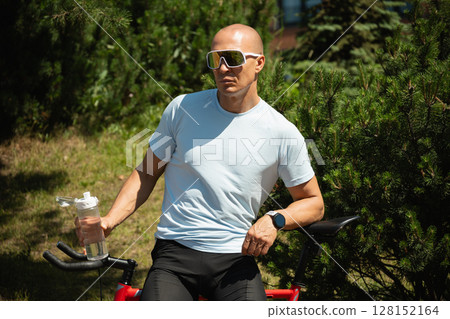 Man in blue t-shirt and sunglasses resting on bicycle holding water bottle at urban public park. Relaxation and refreshment outdoors among greenery. Take a break. Outdoor summer activity. Man in blue t-shirt and sunglasses resting on bicycle holding water bottle at urban public park. Relaxation and refreshment outdoors among greenery. Take a break. Outdoor summer activity. 128152164