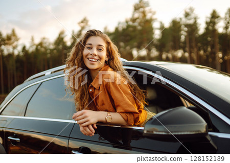 A young woman feels freedom leaning out of a car window in a sunny forest. Travel concept. A young woman feels freedom leaning out of a car window in a sunny forest. Travel concept. 128152189
