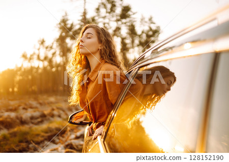 A young woman feels freedom leaning out of a car window in a sunny forest. Travel concept. A young woman feels freedom leaning out of a car window in a sunny forest. Travel concept. 128152190