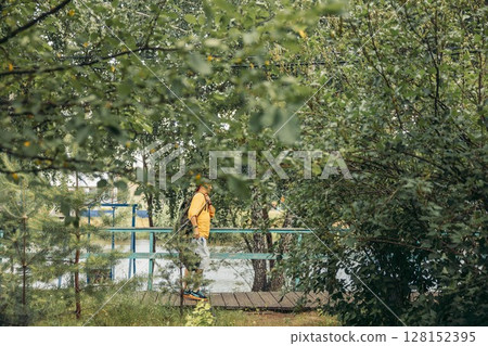 man in orange hoodie standing on small wooden bridge with backpack, lush green foliage and trees near water, eco tourism, travel agency, outdoor adventure company 128152395