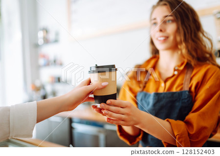 Young woman owner of a coffee shop behind the bar prepares coffee. Concept of orders to go. 128152403