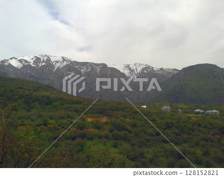 A majestic view of snow-capped mountains towering over a lush green valley with scattered houses and trees under a cloudy sky, showcasing a serene natural scene A majestic view of snow-capped mountains towering over a lush green valley with scattered houses and trees under a cloudy sky, showcasing a serene natural scene 128152821