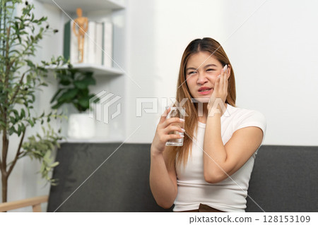 Health Awareness: Young woman holding a glass of water, showing discomfort. Health Awareness: Young woman holding a glass of water, showing discomfort. 128153109