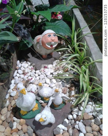 A whimsical garden scene featuring a sheep figurine with a human-like face and several duck figurines, surrounded by lush plants and white pebbles in a decorative outdoor setting A whimsical garden scene featuring a sheep figurine with a human-like face and several duck figurines, surrounded by lush plants and white pebbles in a decorative outdoor setting 128153735