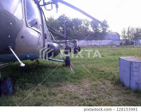 A Military Helicopter with Rocket Pods Parked on a Grassy Field Near Trees and Buildings A Military Helicopter with Rocket Pods Parked on a Grassy Field Near Trees and Buildings 128153991