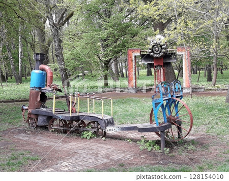 An Artistic Sculpture Resembling a Train with Wheels and a Chimney in a Park Among Trees An Artistic Sculpture Resembling a Train with Wheels and a Chimney in a Park Among Trees 128154010