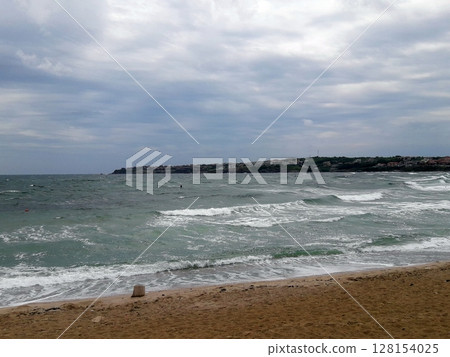 A sandy beach with waves crashing in under a cloudy sky, featuring a distant coastline with buildings and a lone swimmer in the choppy sea waters A sandy beach with waves crashing in under a cloudy sky, featuring a distant coastline with buildings and a lone swimmer in the choppy sea waters 128154025