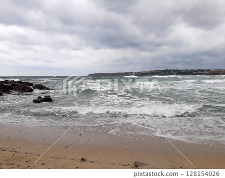 A dramatic seascape featuring turbulent waves crashing against a rocky shoreline under a heavy, overcast sky with a distant coastline visible A dramatic seascape featuring turbulent waves crashing against a rocky shoreline under a heavy, overcast sky with a distant coastline visible 128154026