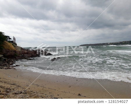 A powerful ocean scene with vigorous waves rolling onto a sandy beach beneath a dark, cloud-filled sky, hinting at an approaching storm A powerful ocean scene with vigorous waves rolling onto a sandy beach beneath a dark, cloud-filled sky, hinting at an approaching storm 128154027