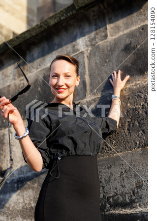 Portrait young adult elegant woman black dress and sunglasses smiles confidently in golden sunlight warm sunset backlit light against historic cathedral building. Female person walking city street 128154090