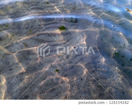 A shallow, clear water surface revealing sandy bottom with gentle ripples and faint underwater details under soft natural lighting A shallow, clear water surface revealing sandy bottom with gentle ripples and faint underwater details under soft natural lighting 128154282