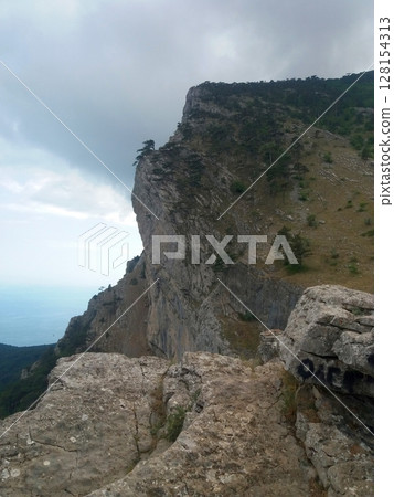 A dramatic cliffside view with rocky terrain, sparse vegetation, and a glimpse of the sea in the distance under a partly cloudy sky A dramatic cliffside view with rocky terrain, sparse vegetation, and a glimpse of the sea in the distance under a partly cloudy sky 128154313