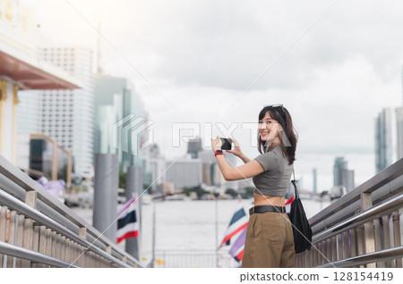 Solo Southeast Asian woman teenager travel blogger and world explorer take photo with smartphone by Chao Phraya river Bangkok Thailand. Asia tourism local cultural experience concept 128154419