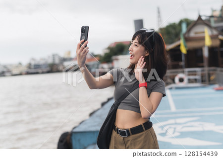 Happy solo southeast asian woman teenager blogger boat. World travel explorer local cultural experience. Asia tourism concept to express joy to friend smartphone and wait Chao Phraya River Bangkok 128154439