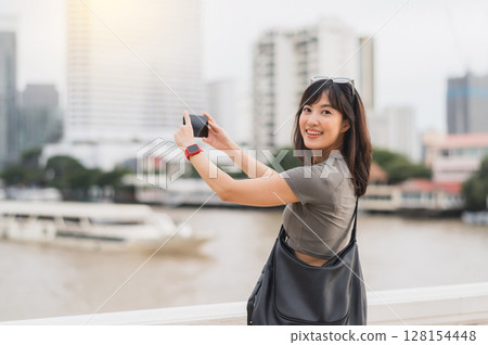 Happy solo Southeast Asian woman teenager blogger on world travel. tourism concept to take photo of boat on Chao Phraya river. local cultural experience with smartphone in Bangkok Thailand 128154448