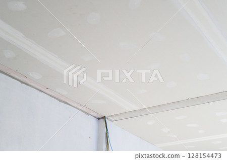 Building interior renovation features gypsum board ceiling installation showing smooth taped edges electrical wire near corner preliminary plastering applied finishing ongoing for new ceilings 128154473