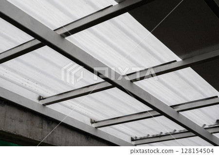 Metal beams supporting translucent sheet roofing. Light filters through corrugated sheet creating patterns. Strong metal framework provides stability shelter overhead. Durable sheet material 128154504