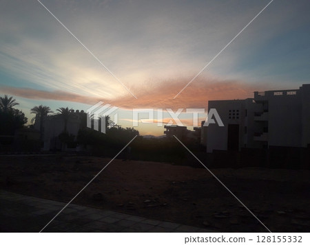 A serene sunset scene with vibrant hues of orange and pink in the sky, silhouetted palm trees, and modern buildings creating a tranquil urban landscape 128155332