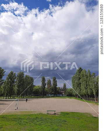 A wide open sports field with a sandy center, framed by tall green trees under a dramatic sky with towering white clouds and hints of blue, creating a striking natural scene A wide open sports field with a sandy center, framed by tall green trees under a dramatic sky with towering white clouds and hints of blue, creating a striking natural scene 128155680