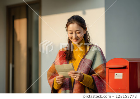 Smiling young woman reading a letter in front of her mailbox 128155778