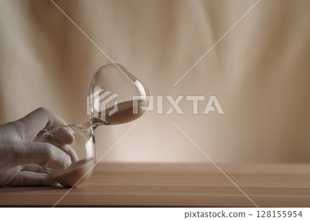 A glove-covered hand holding a tipped hourglass on a wooden surface, symbolizing the flow of time, creativity, and patience in a contemporary minimalist style. 128155954