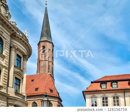 Historic Gothic Church Tower of Heilig-Geist in Munich: Red Tiled Roof and Medieval Brick Architecture Framed by Ornate Facade and Blue Sky in the Bavarian Old Town 128156739