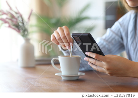 Woman hand using phone stirring coffee at home Woman hand using phone stirring coffee at home 128157019