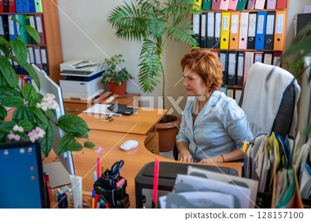 Woman working at a cluttered office desk with folders, documents, and plants in the background. Concept of everyday workflow, paperwork routine, and real administrative work environment Woman working at a cluttered office desk with folders, documents, and plants in the background. Concept of everyday workflow, paperwork routine, and real administrative work environment 128157100