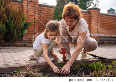 Woman gardening with a young girl in the backyard, teaching her how to care for flowers. Family bonding, outdoor learning, and eco-friendly lifestyle. Woman gardening with a young girl in the backyard, teaching her how to care for flowers. Family bonding, outdoor learning, and eco-friendly lifestyle. 128157105