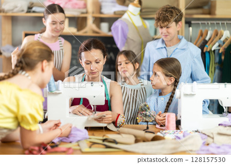 Woman teacher demonstrates how to sew on a sewing machine. Children gather around and watch 128157530