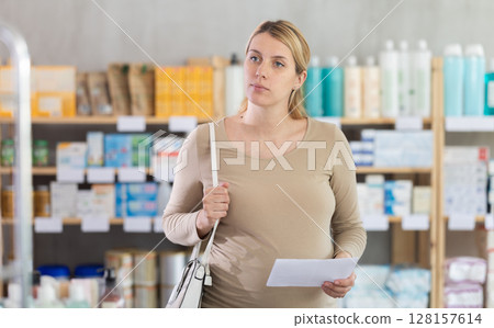 Pregnant woman with doctors prescription is looking for vitamins on pharmacy display case 128157614